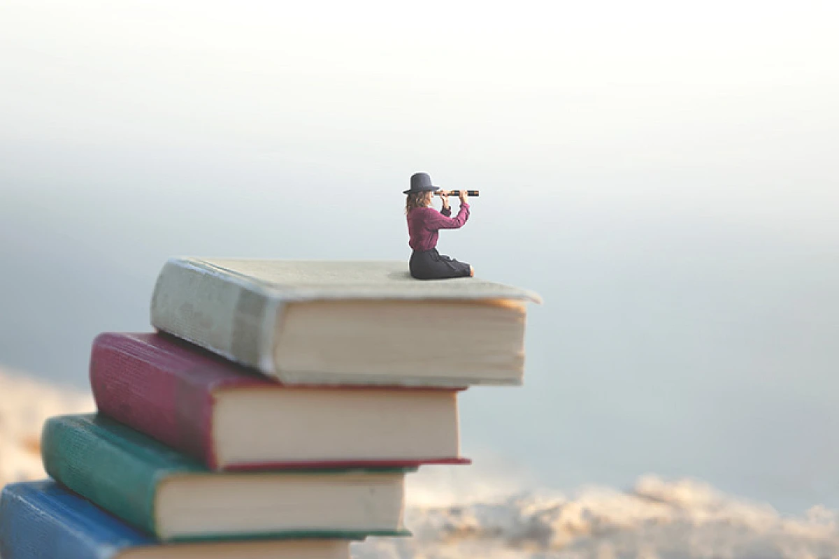 Modellfigur mit Fernglas sitzt auf einem Stapel Bücher und sieht in die Ferne.