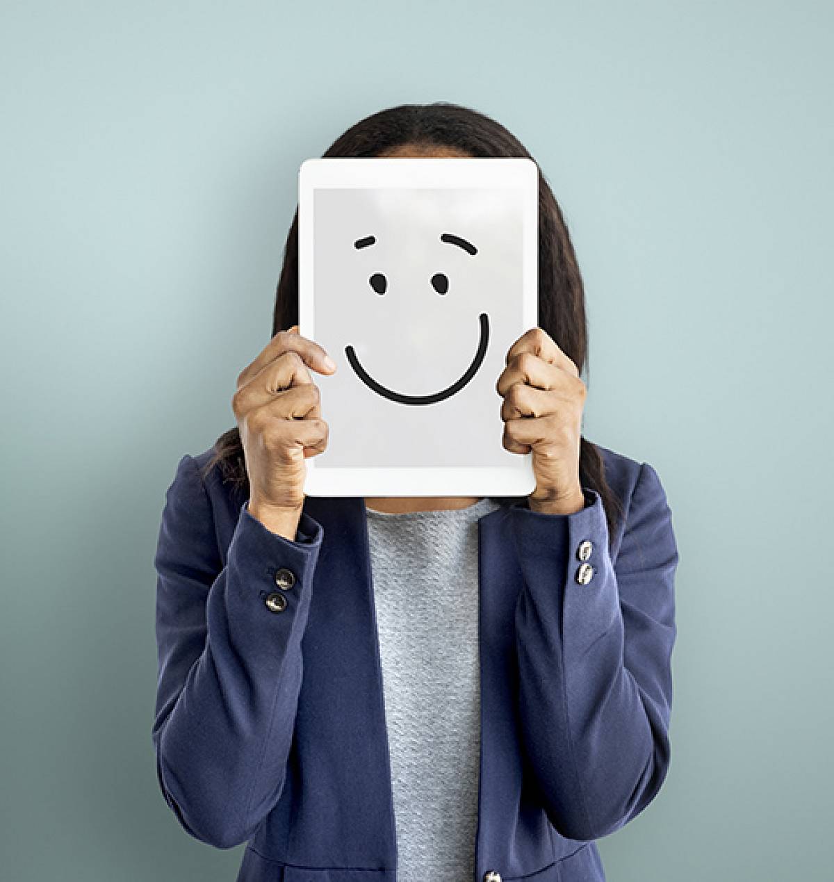 Frau im Blazer hält sich ein Tablet vors Gesicht, auf dem ein lachender Smiley zu sehen ist.
