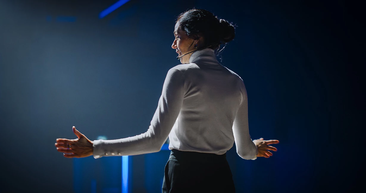 Frau mit weißer Bluse auf einer Bühne, die im Scheinwerferlicht eine Rede hält.