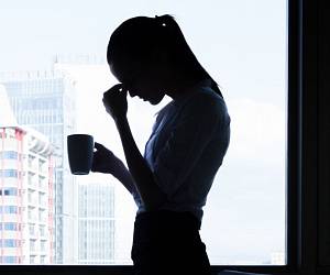 Silhouette einer gestressten Frau steht mit Kaffeetasse in der Hand vor einem Fenster, während sie sich mit der Hand an die Stirn fasst.