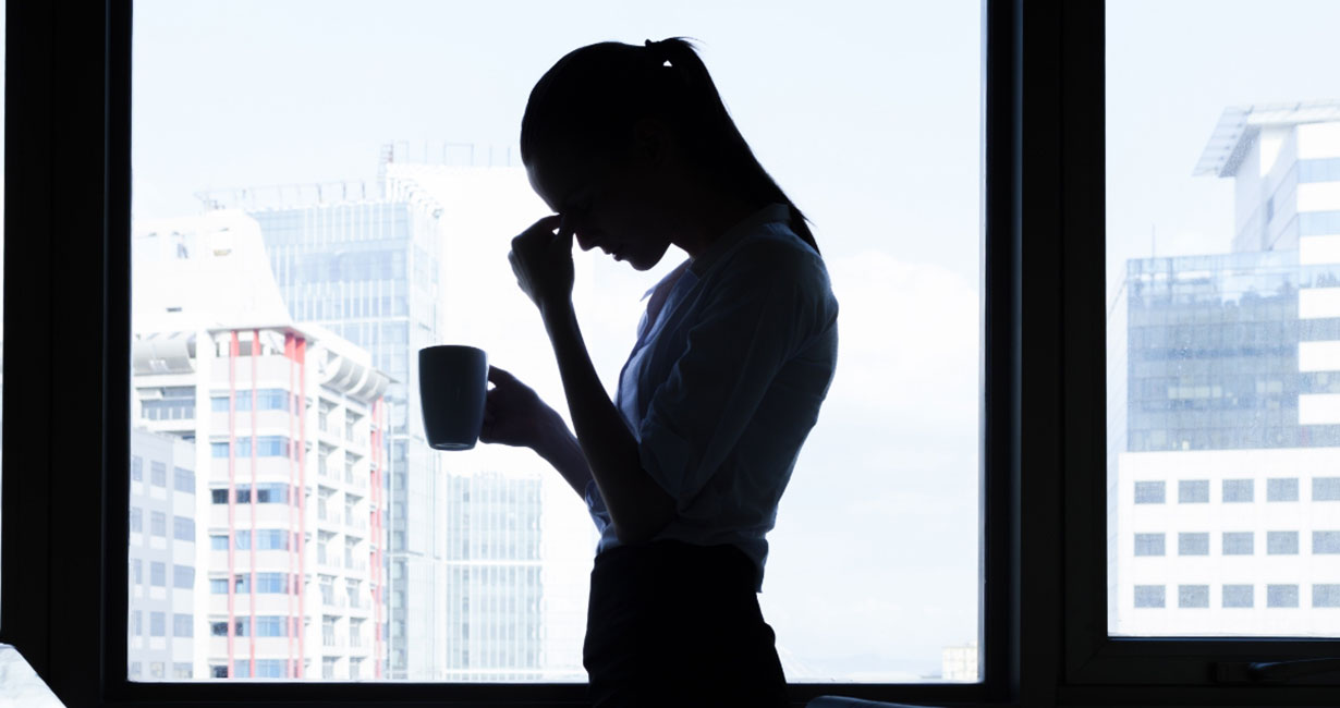 Silhouette einer gestressten Frau steht mit Kaffeetasse in der Hand vor einem Fenster, während sie sich mit der Hand an die Stirn fasst.