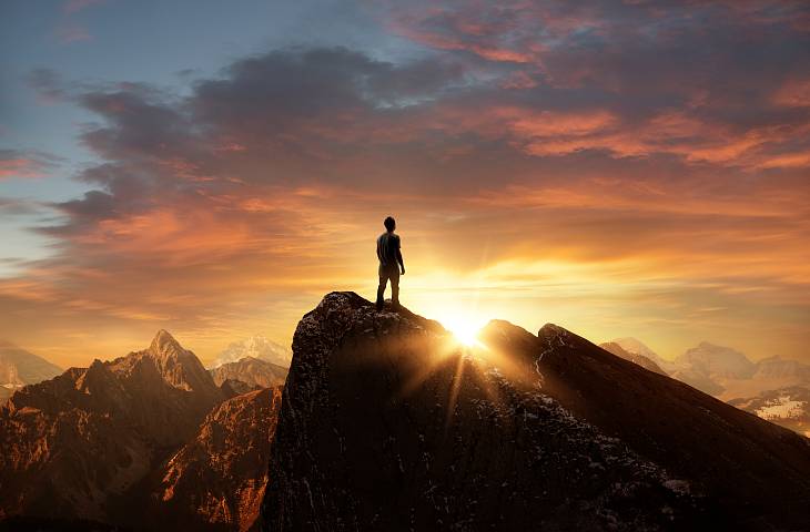 Mann steht auf einer Bergspitze und blickt Richtung Horizont entgegen der Sonne