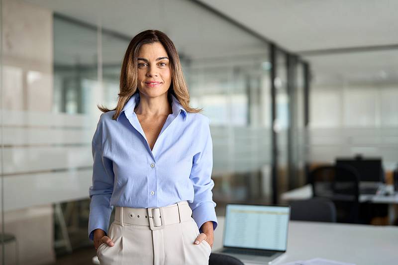 Junge brünette Frau in blauer Bluse und heller Hose steht in einem Besprechungsraum und sieht gespannt in die Kamera.
