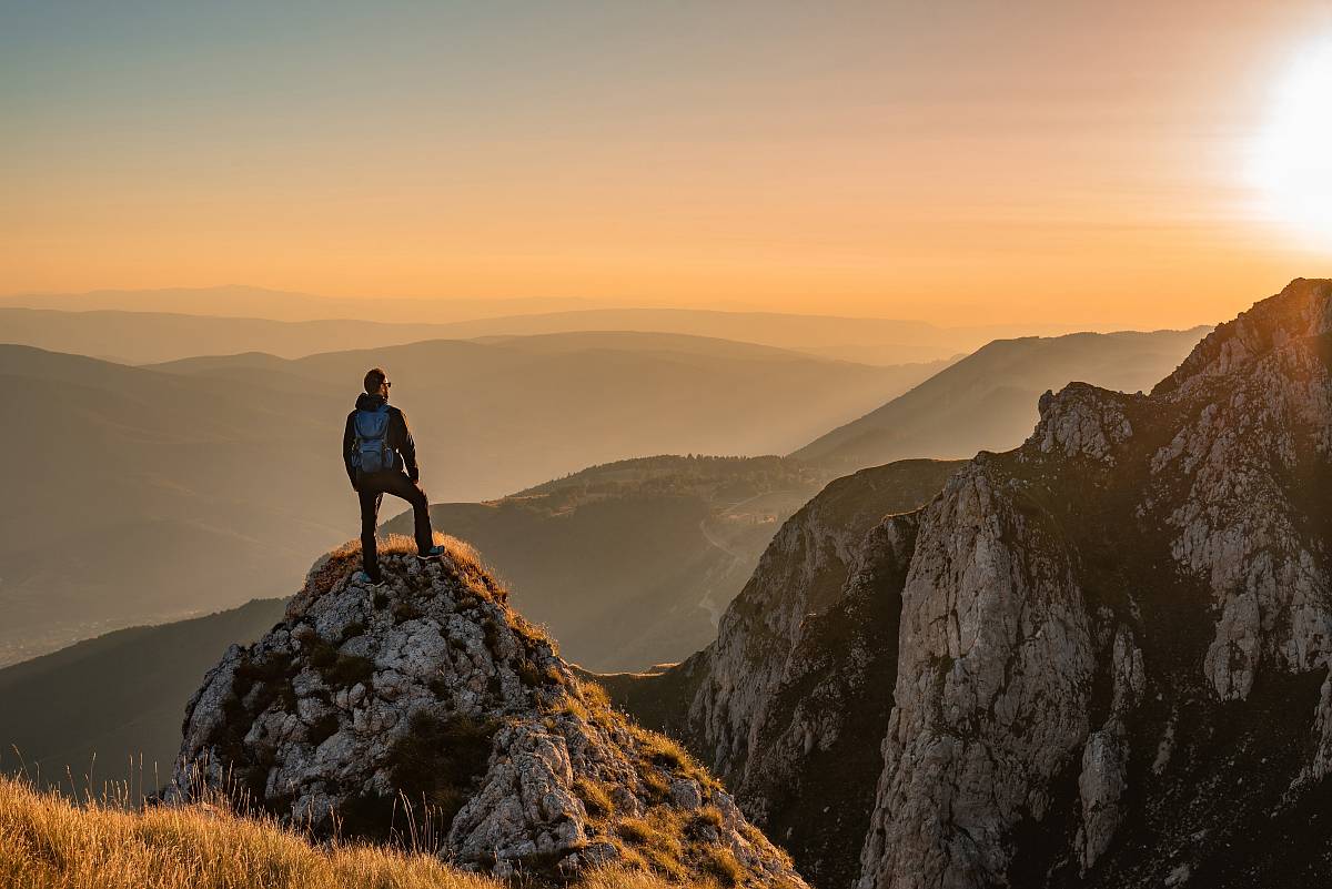 Ein Wanderer mit Rucksack steht auf einem Felsen und blickt bei Sonnenuntergang auf ein weites, bergiges Tal.