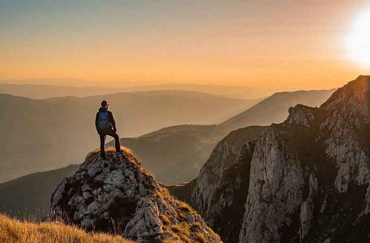 Ein Wanderer mit Rucksack steht auf einem Felsen und blickt bei Sonnenuntergang auf ein weites, bergiges Tal.