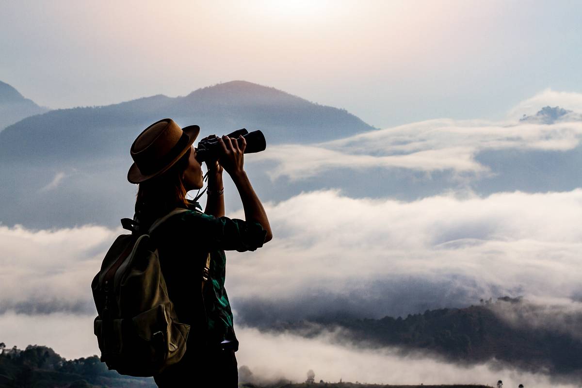 Eine Frau mit Hut und Rucksack blickt durch ein Fernglas auf eine nebelige Berglandschaft.