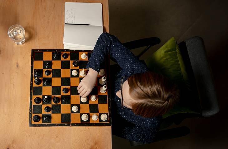 Eine Person spielt Schach an einem Tisch, neben dem ein Glas Wasser und ein Notizbuch liegen.