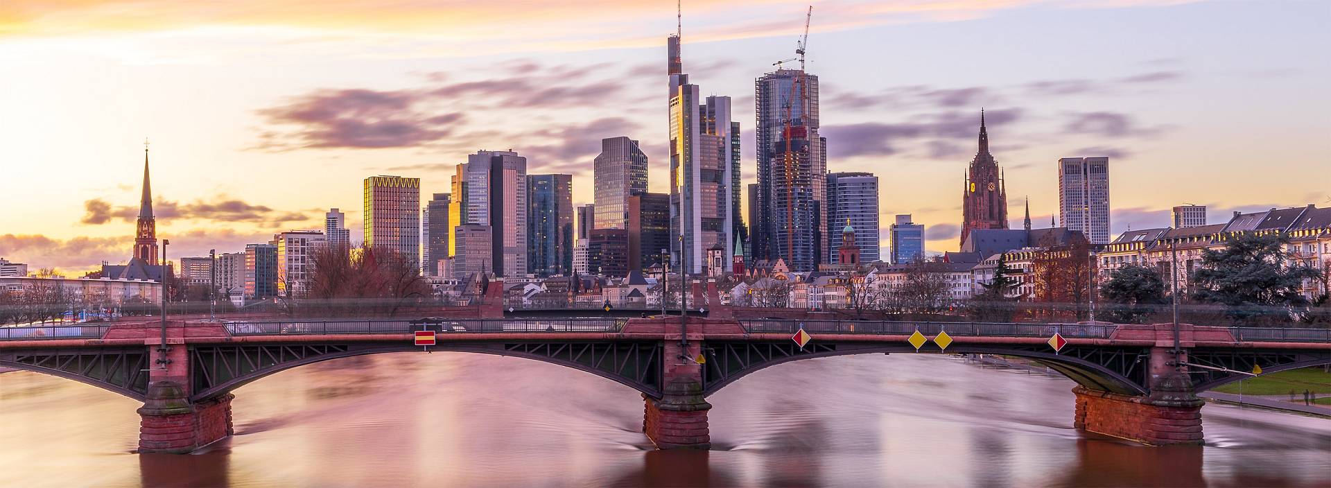 Skyline von Frankfurt am Main bei Sonnenuntergang, mit moderner Architektur, Brücke über den Main und historischer Kirche im Vordergrund.