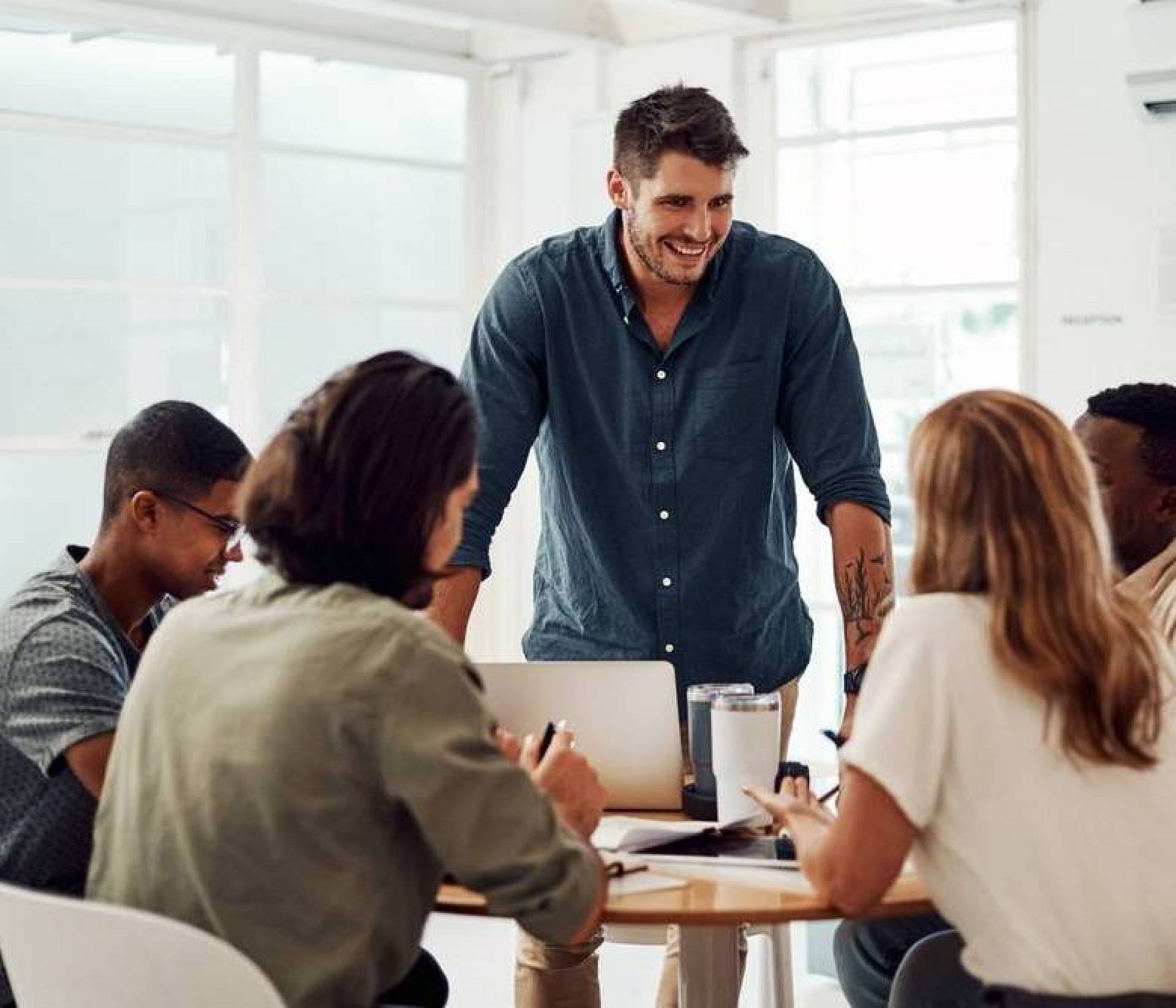 Gruppe junger Berufstätiger bei einem Meeting, ein Mann steht lächelnd am Tisch und führt die Besprechung an.