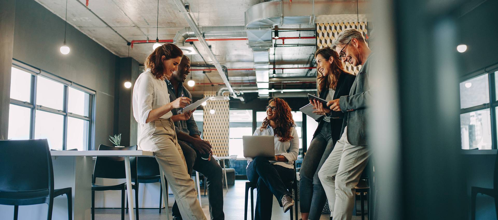Teammeeting in einem modernen Büro, mehrere Personen stehen und sitzen gemeinsam mit Laptop und Smartphones