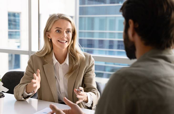 Eine Frau im Business-Outfit sitzt an einem Tisch in einem modernen Büro und spricht gestikulierend mit einem Mann, der ihr gegenübersitzt. Beide befinden sich in einem Gespräch vor großen Fenstern mit Blick auf Bürogebäude.
