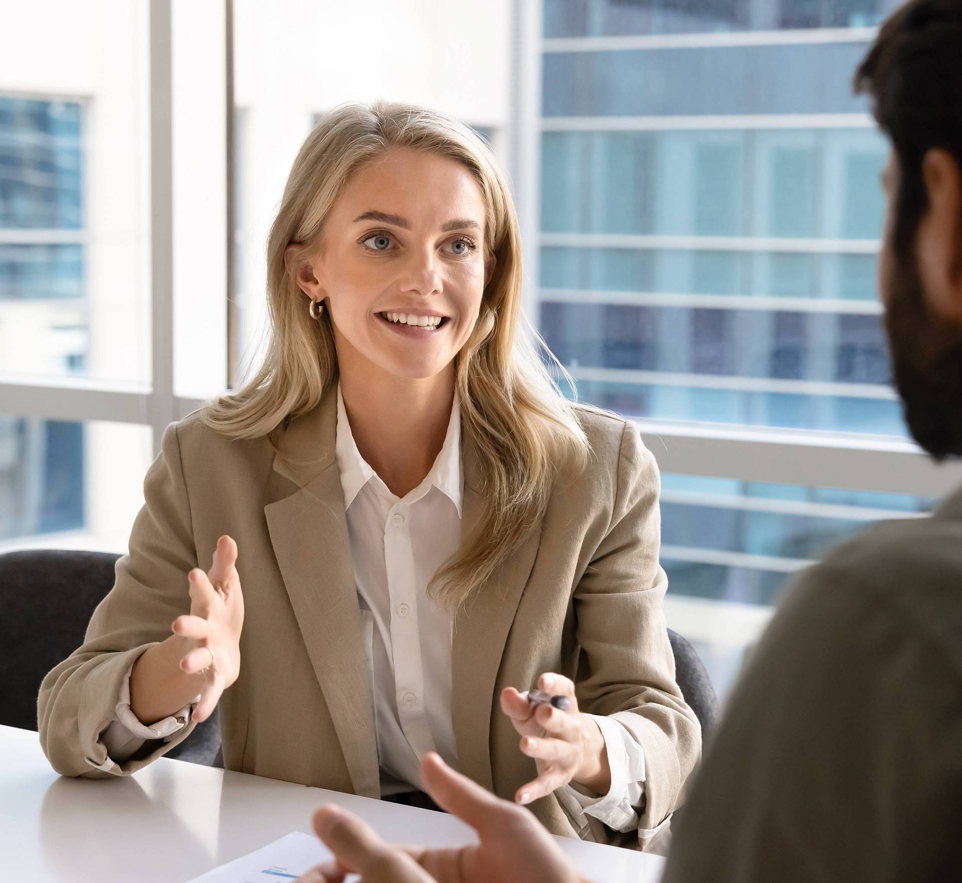 Eine Frau im Business-Outfit sitzt an einem Tisch in einem modernen Büro und spricht gestikulierend mit einem Mann, der ihr gegenübersitzt. Beide befinden sich in einem Gespräch vor großen Fenstern mit Blick auf Bürogebäude.