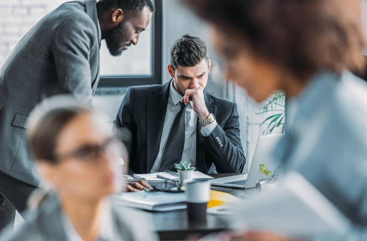 Nachdenkliche Geschäftspersonen in einem Meeting, während ein Kollege am Tisch konzentriert auf Unterlagen blickt.