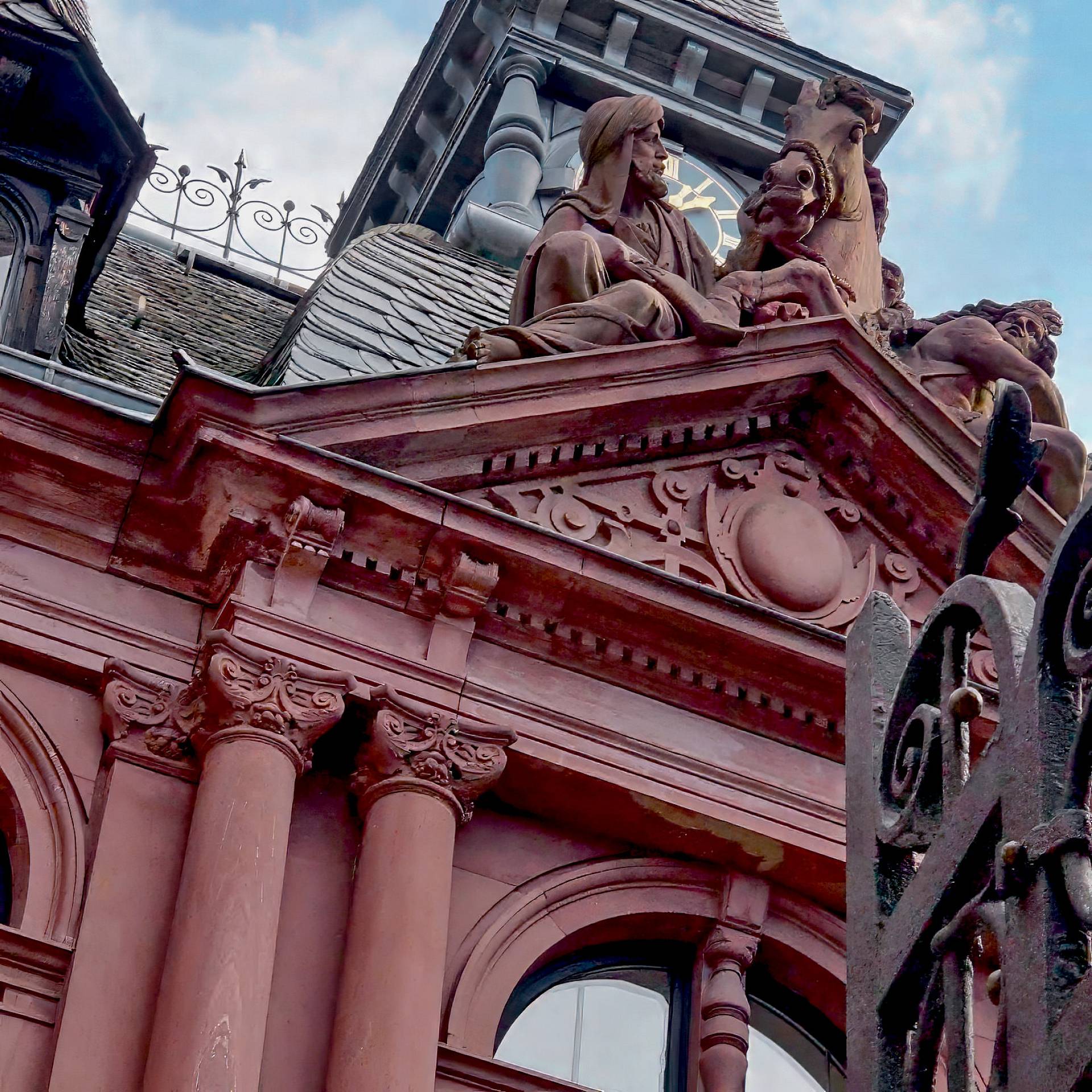 Detailaufnahme der historischen Fassade des Frankfurter PresseClubs mit rotem Sandstein, Säulen und figürlichen Skulpturen auf dem Dachgiebel vor blauem Himmel